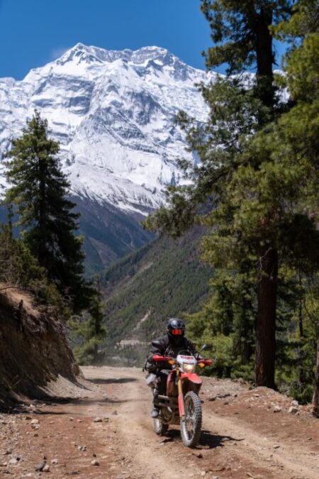 An adventure rider on dual-sport motorcycles navigate a rugged dirt trail in Manang, with the massive, snow-capped peaks of the Annapurna Himalayan range towering in the backdrop.